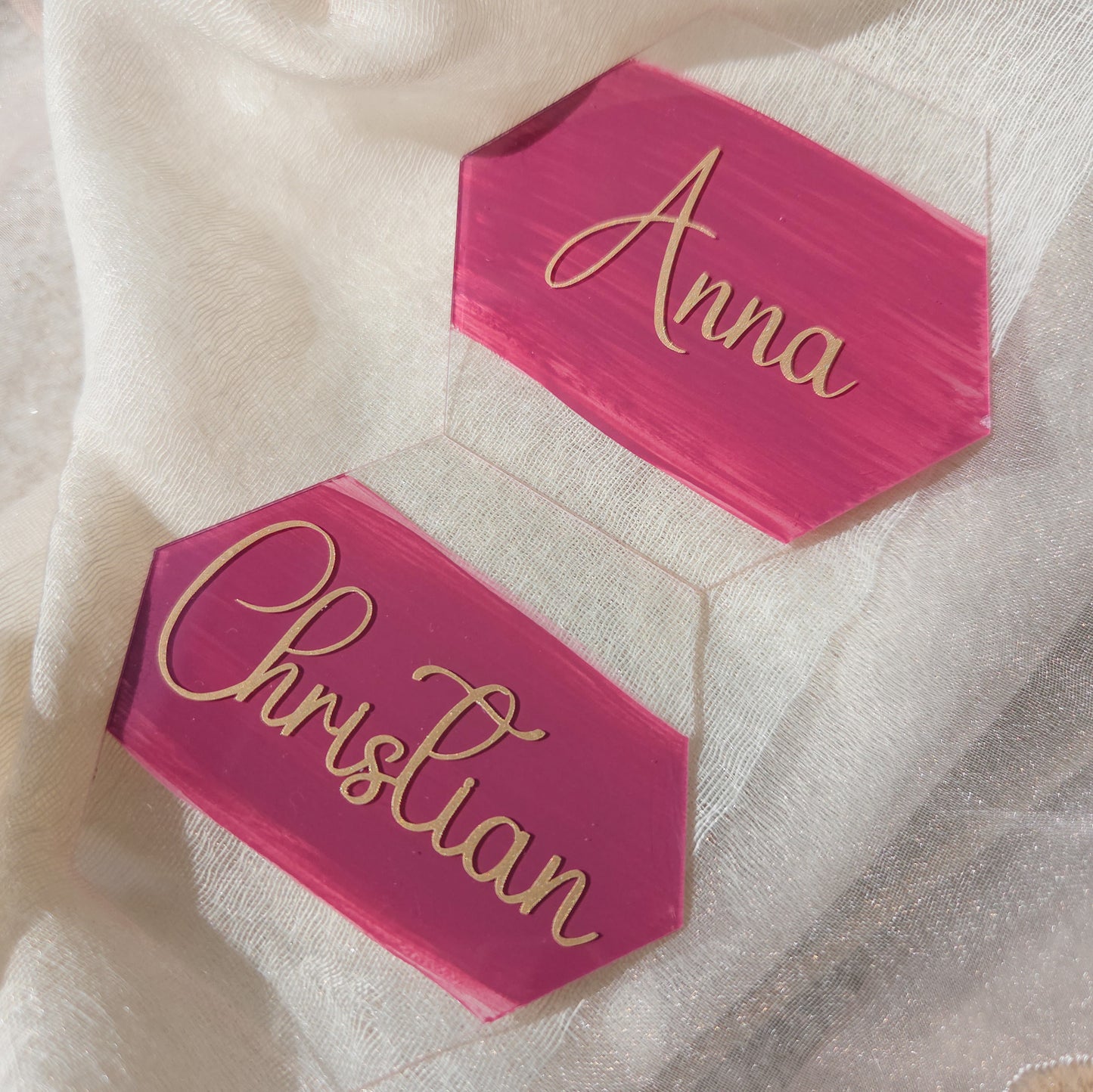 Personalisierte pinke Acryl-Namensschilder mit goldener Schrift für Anna und Christian auf hellem Stoffhintergrund als stilvolle Tischkarten.