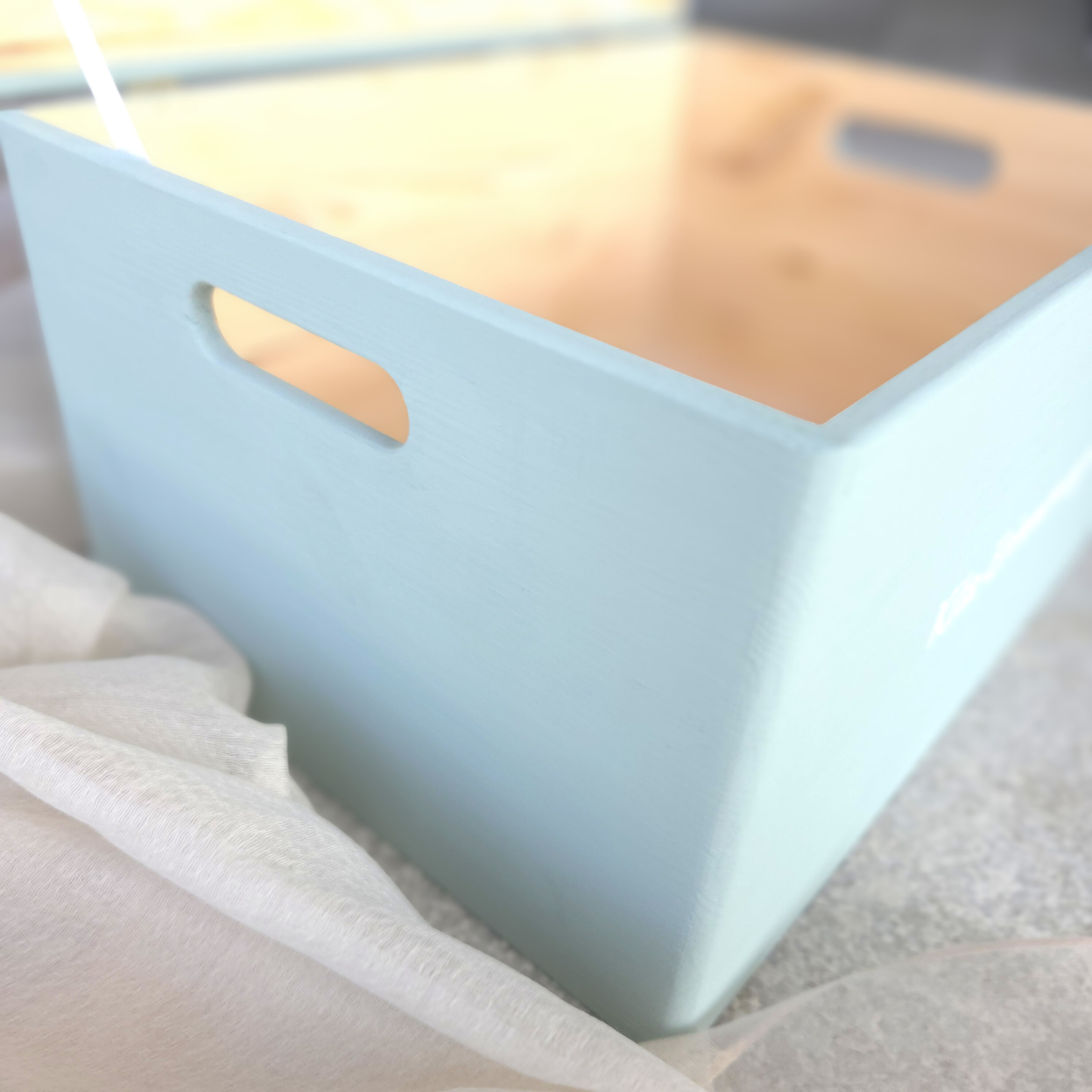 Hellblaue Kita-Erinnerungsbox aus Holz, seitlich fotografiert mit ausgeschnittenem Tragegriff und hellem Stoff im Hintergrund, zeigt stabile Verarbeitung und grosses Fassungsvermögen.