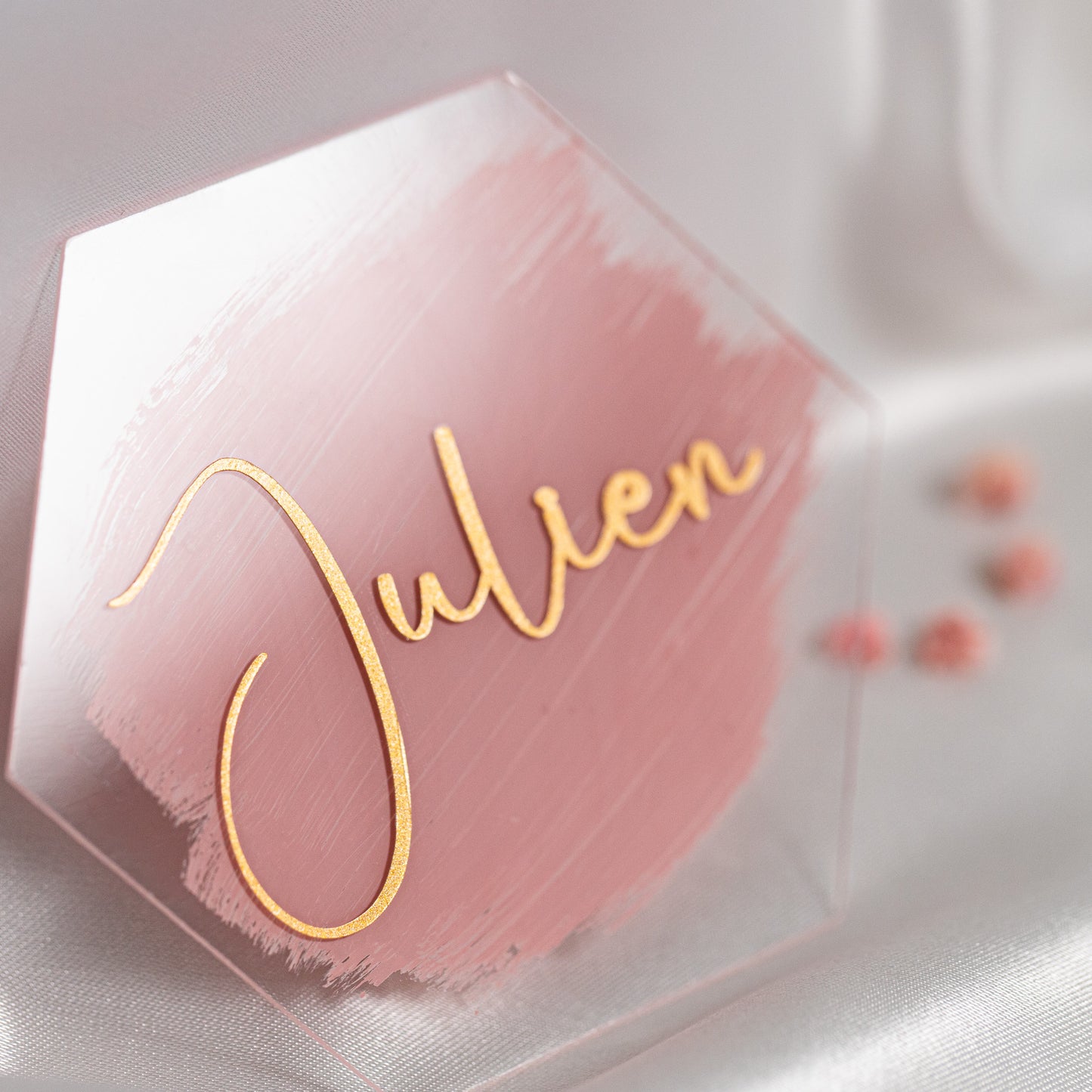 Hexagonales Acryl-Namensschild mit rosa Brush-Hintergrund und goldener Handschrift „Julien“, stilvoll arrangiert auf weissem Satin als elegante Platzkarte für die Hochzeit.
