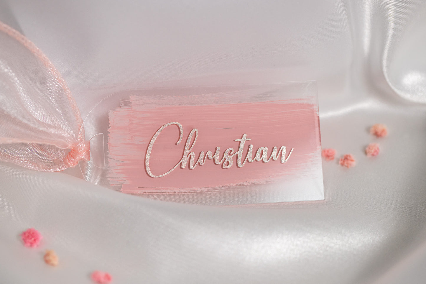 Rechteckiges Acryl-Namensschild mit rosa Brush-Hintergrund und glitzernder Schrift „Christian“, dekoriert auf weissem Satin mit zarten rosa Blüten als elegante Platzkarte für die Hochzeit.