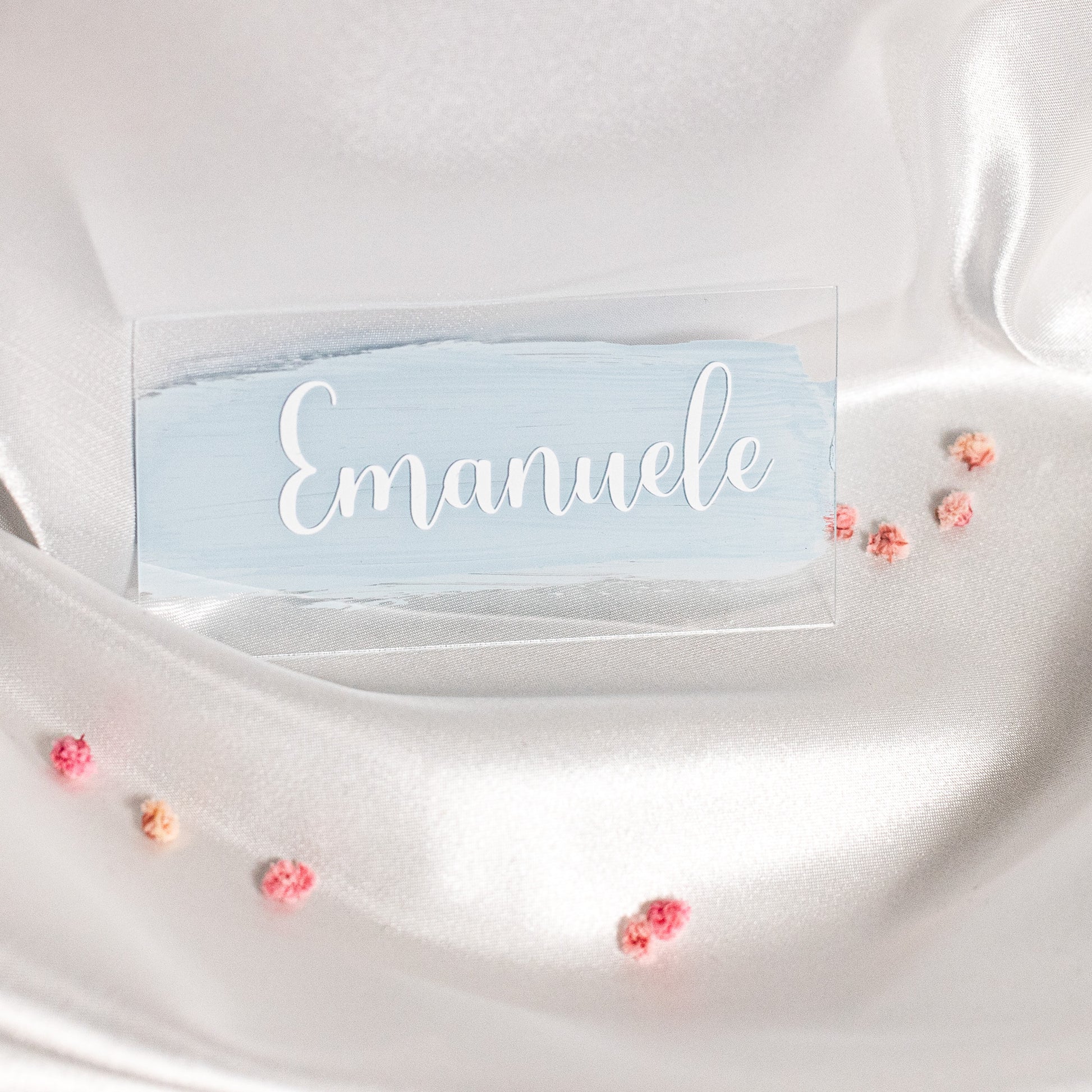 Rechteckiges Acryl-Namensschild mit hellblauem Brush-Hintergrund und weisser Schrift „Emanuele“, dekorativ auf weissem Satin mit kleinen rosa Blüten arrangiert.