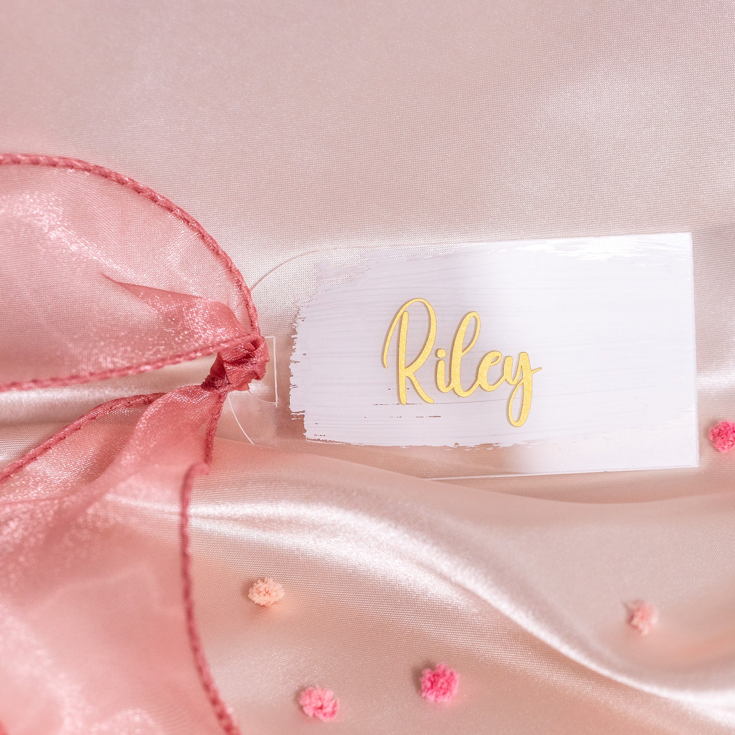 Kleines rechteckiges Acryl-Namensschild mit weissem Brush-Hintergrund und goldener Schrift „Riley“, dekoriert mit rosa Organzaschleife auf Satin als elegante Platzkarte für die Hochzeit.