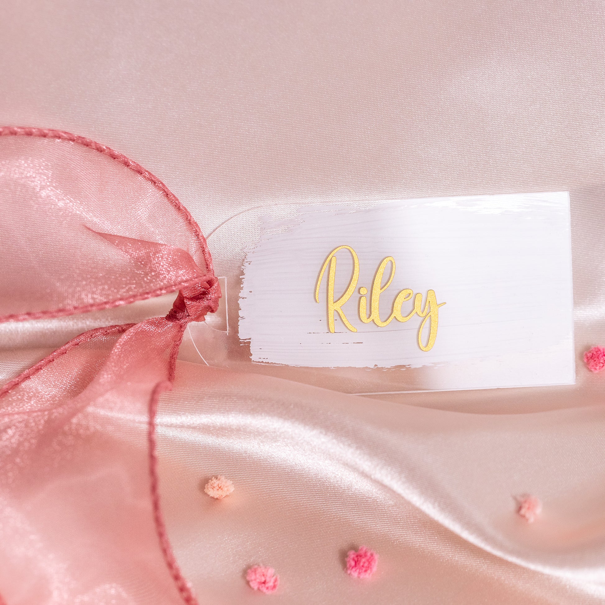 Kleines rechteckiges Acryl-Namensschild mit weissem Brush-Hintergrund und goldener Schrift „Riley“, dekoriert mit rosa Organzaschleife auf Satin als elegante Platzkarte für die Hochzeit.