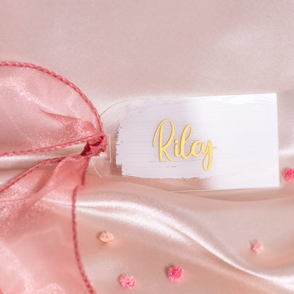 Kleines rechteckiges Acryl-Namensschild mit weissem Brush-Hintergrund und goldener Schrift „Riley“, dekoriert mit rosa Organzaschleife auf Satin als elegante Platzkarte für die Hochzeit.